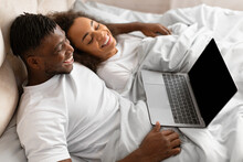 Happy young black couple relaxing in bed using laptop indoor