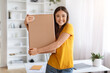 © Prostock-studio - Cheerful asian woman holding cardboard box while standing near desk at home