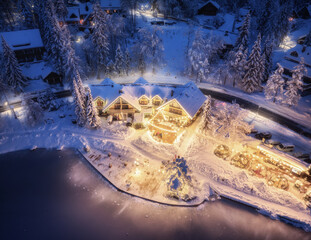 Wall Mural - Aerial view of illuminated houses in fairy village in snow, forest, Jasna lake, street lights at winter night. Top view of alpine countryside, snowy pine trees at dusk. Kranjska Gora, Slovenia