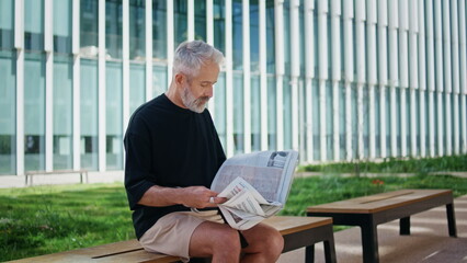 Senior person reading newspaper on street. Good looking mature man resting bench