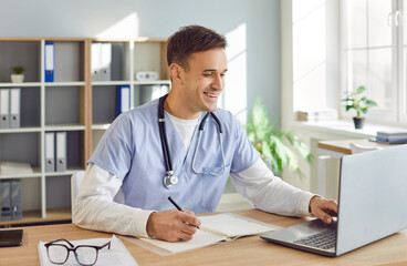 Portrait of a young male doctor or nurse in blue medical uniform sitting at the desk and working in office. Man physician wearing stethoscope in clinic using laptop typing and writing in notepad.