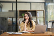 © Wasana - Attractive Asian businesswoman sitting with laptop and documents at table in office, thinking an ideas.