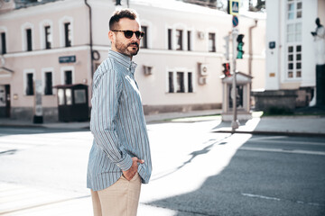 Wall Mural - Portrait of handsome confident stylish hipster lambersexual model. Sexy modern man dressed in blue shirt and trousers. Fashion male posing on street background in Europe city at sunset. In sunglasses