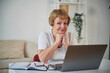 © standret - Front view. Sitting by table with laptop. Senior elderly woman is at home in the living room