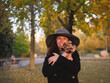 Beautiful stylish elegant joyful woman in a large hat and black coat portrait with a small Yorkie dog in her arms