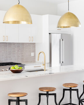 A kitchen detail with white cabinets, gold faucet and light hanging over the island with bar stools, and a tiled backsplash.