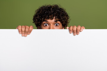 Photo of impressed scared guy rising hiding behind white poster empty space isolated khaki color background