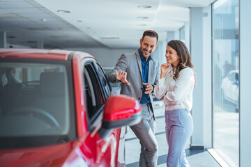 Wall Mural - Beautiful young couple at car showroom choosing a new car to buy. Happy beautiful couple is choosing a new car at dealership. Happy young couple standing alongside their dream car and looking in