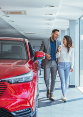 Wall Mural - Beautiful young couple at car showroom choosing a new car to buy. Happy beautiful couple is choosing a new car at dealership. Happy young couple standing alongside their dream car and looking in