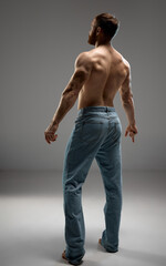 Rear view portrait of strong male model with healthy back posing in denim jeans against grey studio background. Men's skincare promotion focusing on natural body care. Concept of beauty and fashion.