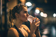 © arhendrix - a white female gym athlete drinking cold isotonic sports water drink. sweaty after exercises. blurry gym background.