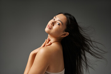 Wall Mural - pretty asian woman with acne prone skin and shiny brunette hair waving on wind on grey background