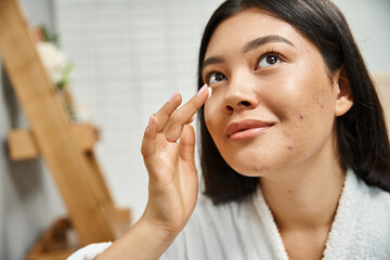 Wall Mural - brunette asian woman with acne applying cream on face and looking up in bathroom, skin issues