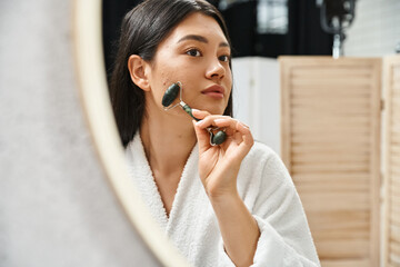 Wall Mural - young asian woman with brunette hair using jade roller for a facial massage in bathroom, skin care