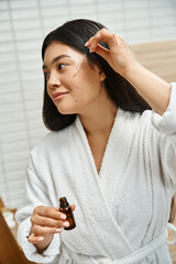 Wall Mural - smiling and young asian woman in bath robe applying facial serum to treat acne on face near mirror