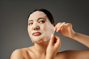 Wall Mural - beautiful young asian woman applying hydrating sheet mask for skin treatment on grey background