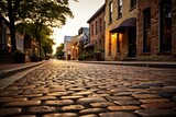 Fototapeta Uliczki -  a cobblestone street with buildings and trees in the background at sunset in a small town with cobblestones on both sides of the street and a cobblestones on both sides.