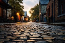  A Cobblestone Street In A Small Town At Dusk With The Sun Shining On The Buildings And The Cobblestones On The Street In Front Of The Street.