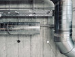 The image shows a metal ventilation duct system on a concrete wall. It has two cylindrical ducts connected at an elbow joint. The wall has some holes and objects on it.