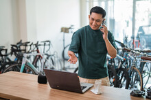 young man makes a cell phone call while working on a laptop at a desk inside a bicycle shop