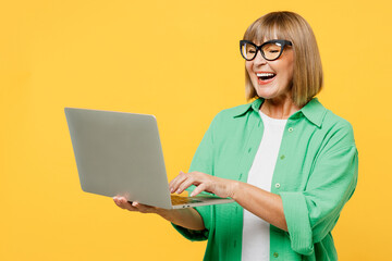 Wall Mural - Elderly smiling happy blonde IT woman 50s years old wear green shirt glasses casual clothes hold use work on laptop pc computer isolated on plain yellow background studio portrait. Lifestyle concept.