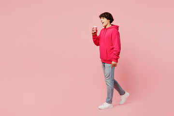 Full body side view young Caucasian man wears hoody casual clothes hold takeaway delivery craft paper brown cup coffee to go isolated on plain pastel light pink background studio. Lifestyle concept.