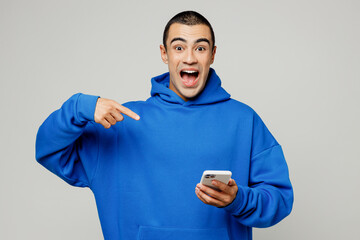 Wall Mural - Young surprised middle eastern man wear blue hoody casual clothes hold in hand use point index finger on mobile cell phone isolated on plain solid white background studio portrait. Lifestyle concept.