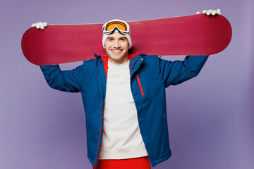 Wall Mural - Smiling happy man he wear warm blue windbreaker jacket ski goggles mask hat hold snowboard on shoulder look camera spend extreme weekend winter season in mountains isolated on plain purple background