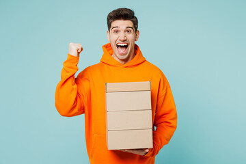 Wall Mural - Young excited man he wears orange hoody casual clothes hold stack cardboard blank boxes do winner gesture isolated on plain pastel light blue cyan color background studio portrait. Lifestyle concept.