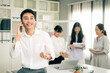 © Wanwajee - Young Asian business man using smartphone while sitting on table in office with smile. Man in white shirt looking away while talking to someone on mobilephone with co-worker background.