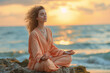 Young woman dressed in peach color meditating on a rock by the sea at sunset to improve her mental health.