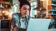 A stylish woman with bold tattoos on her arm sits indoors, gazing intently at her laptop, her fashionable clothing adding to the overall aesthetic of the scene