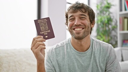 Poster - Confident bearded young man sitting on his living room sofa, grinning wide with czech republic passport, overflowing with happiness, ready to embark on an exciting vacation