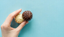 Young Adult Woman Hand Fingers Holding Fresh And Old Spoiled Dark Chocolate Candies On Light Blue Table Background. Pastel Color. Compare Two Pralines. Point Of View Shot. Top Down View