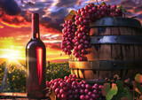 Fototapeta  - Bottle of white wine beside wooden barrel, grapes against vineyard sunset