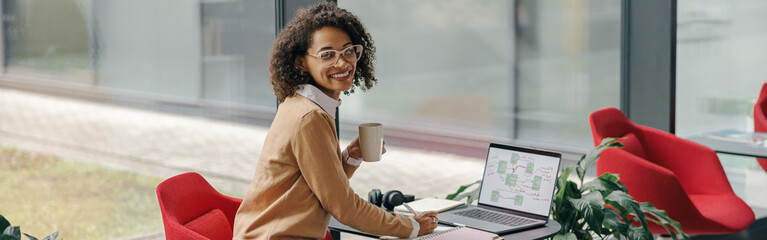 Wall Mural - Smiling female freelancer have coffee break during working on laptop in modern coworking