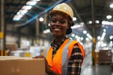 Fototapeta  - A smiling dark-skinned female worker wearing an orange vest and safety helmet and holding a package in a modern warehouse