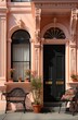 A charming pink building adorned with black doors and chairs, with lush houseplants and vibrant flowerpots lining the outdoor windows and molding, creating a cozy and inviting home