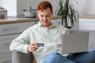 Canvas Print - Handsome young man with credit card and modern laptop sitting on sofa while shopping online