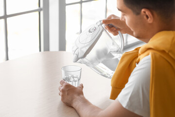 Wall Mural - Young man pouring purified water into glass from filter jug at table in kitchen