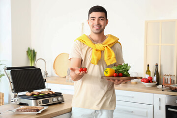 Canvas Print - Young man with board of fresh vegetables cooking tasty sausages on modern electric grill in kitchen