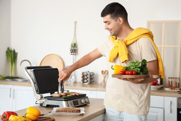 Canvas Print - Young man with board of fresh vegetables cooking tasty sausages on modern electric grill in kitchen