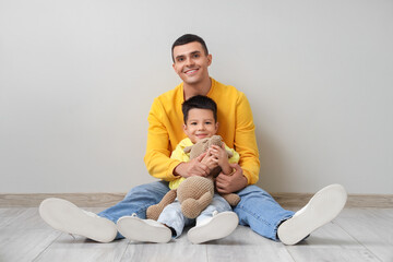 Poster - Young man and his little son with toy bear sitting near light wall