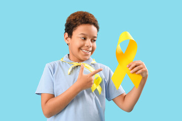 Poster - Cute little African-American boy in superhero costume pointing at yellow ribbon on blue background. Childhood cancer awareness concept