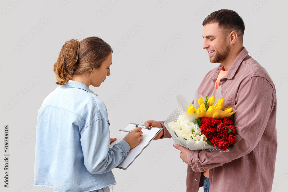 Young woman signing documents after receiving flowers from courier on light background