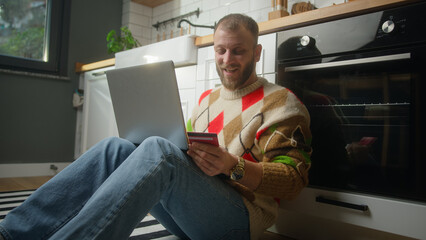 Man sitting on the floor in kitchen entering credit card number on laptop computer for makes distant goods purchase. Credit card is declined or out of limit, feeling sad and unhappy