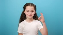 Portrait Of Adorable Smiling Little Girl Child Showing Ok Gesture To Camera, Expression Optimism, Wearing White Casual T-shirt, Posing Isolated Over Plain Blue Color Background Wall In Studio