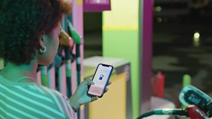 Over shoulder footage of young modern Black woman using gas refueling app processing digital refueling at self-service petrol station at night