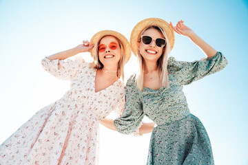 Wall Mural - Two young beautiful smiling hipster woman in trendy summer dress. Carefree women in the street in hats. Positive models at sunset. Cheerful and happy. Bottom view. They look at camera from above