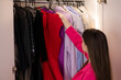 Side view, no face. Cute young woman standing in front of a coat rack and trying to choose an outfit for a date, birthday, party. Wardrobe selection, stylish fashionable clothes, stylist, shopping